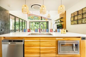 A modern kitchen with wooden cabinets, a built-in dishwasher, a microwave, and a large window overlooking greenery. - Villa Bosque (La Fortuna)