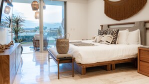 Ein Schlafzimmer mit einem großen Bett, einem Holznachttisch, einem Beistelltisch, einer Hängeleuchte und Blick ins Freie.