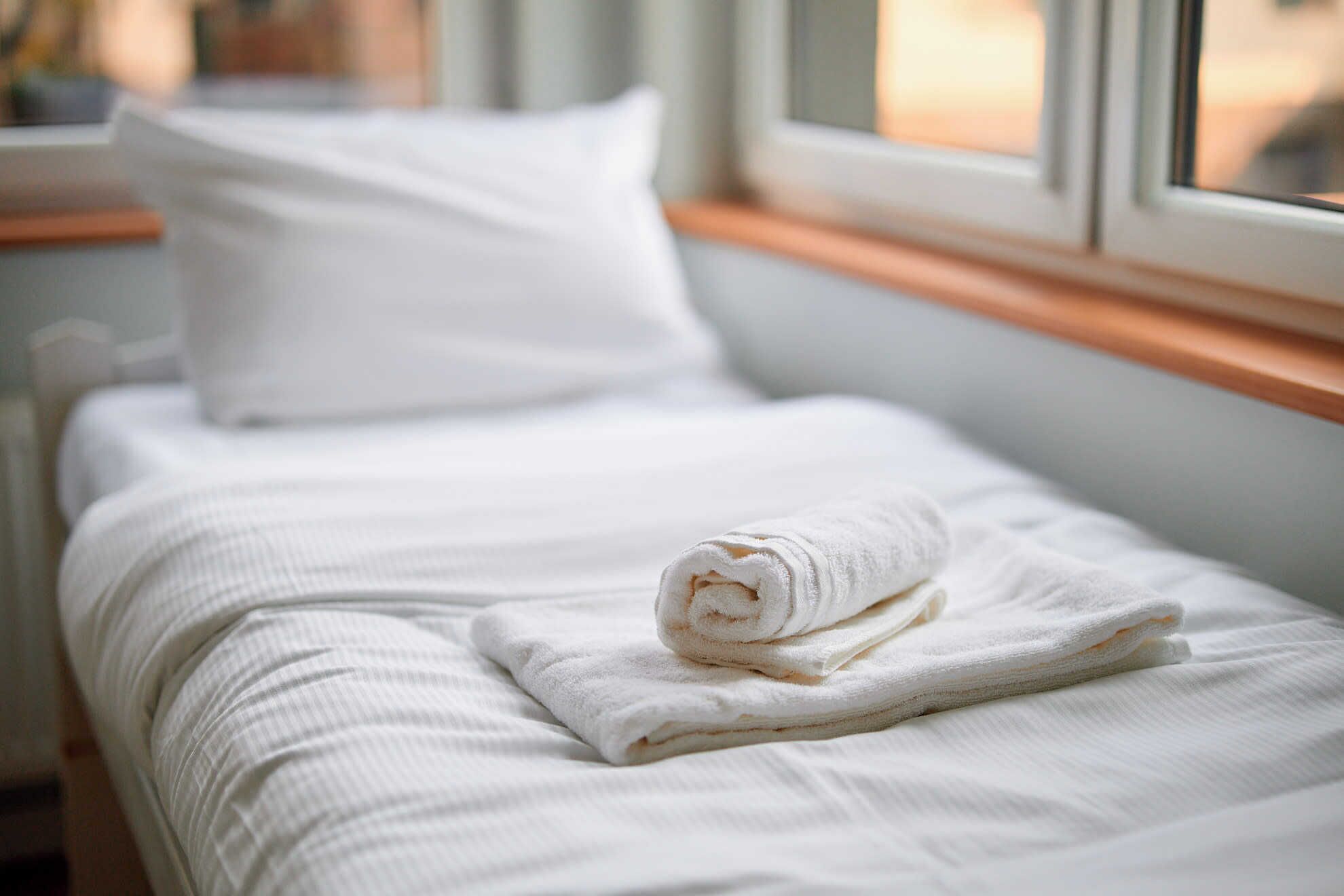 Una cama bien hecha con una toalla enrollada, ropa de cama blanca y una ventana con marco de madera.