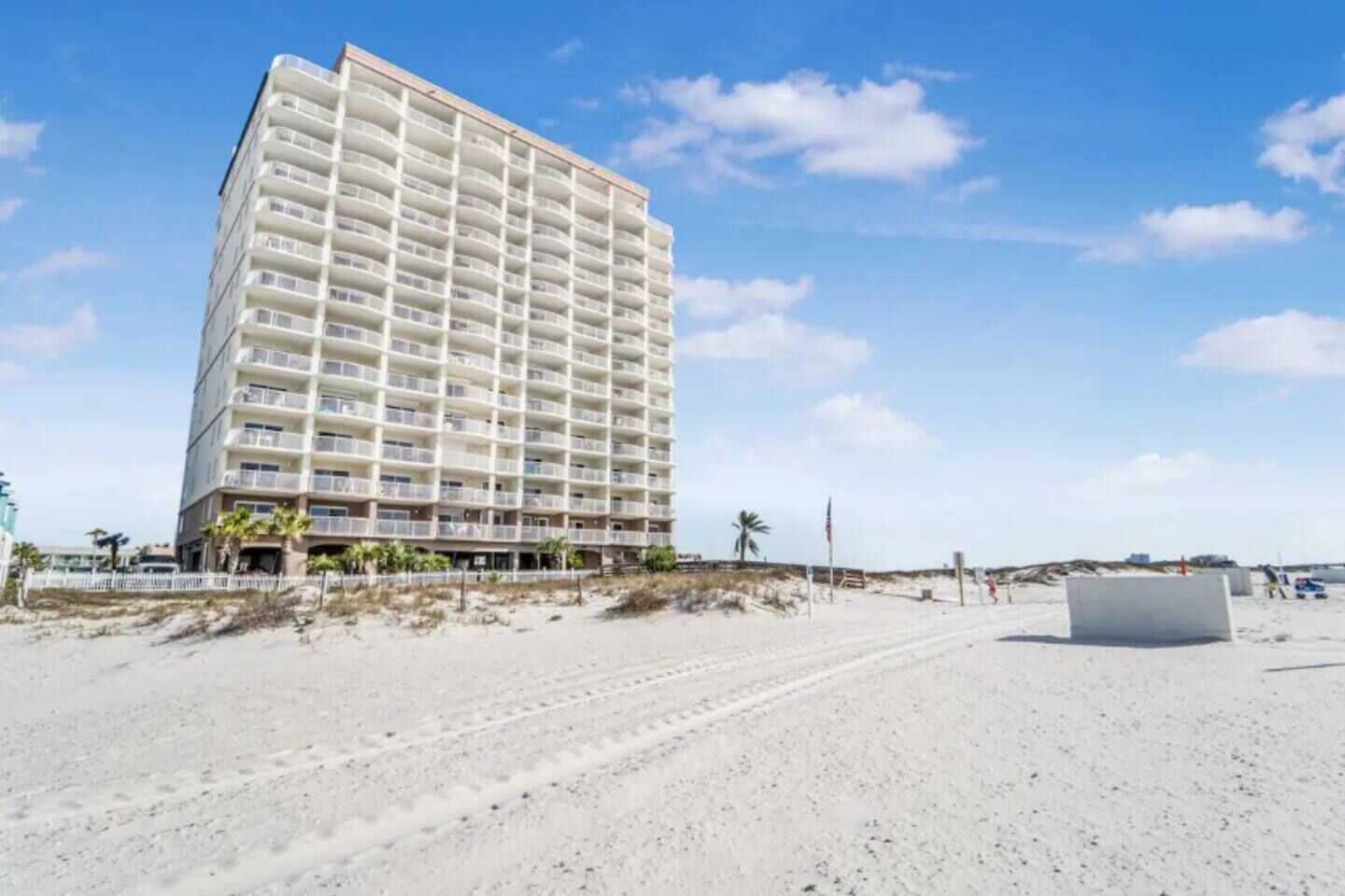 A tall, multi-story hotel with balconies, located on a sandy beach with a clear sky.