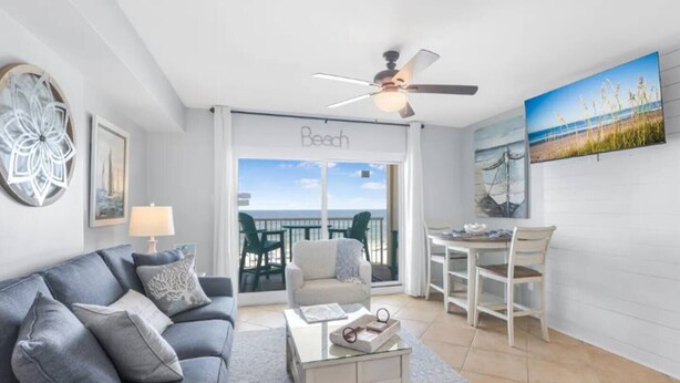 Een woonkamer met een grijze bank, een salontafel en een plafondventilator. Er is een balkon met uitzicht op het strand.