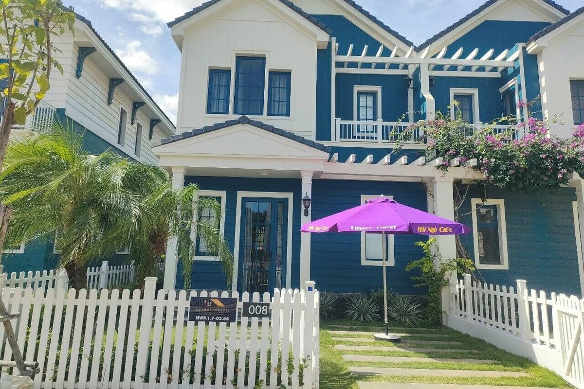 A two-story building with a white picket fence, a purple umbrella, and a sign indicating a luxury travel destination.