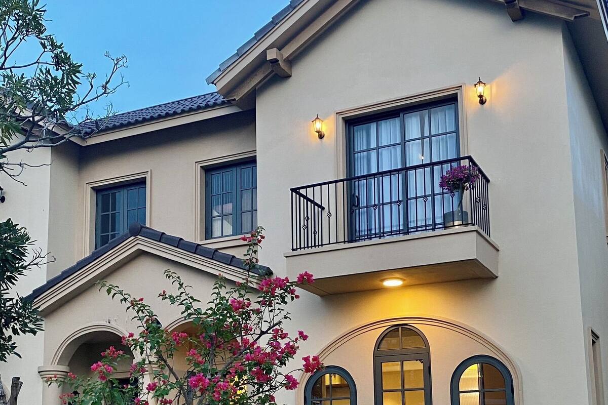 A two-story house with a white picket fence, a balcony with black railings, and a small tree with pink flowers in the front yard.