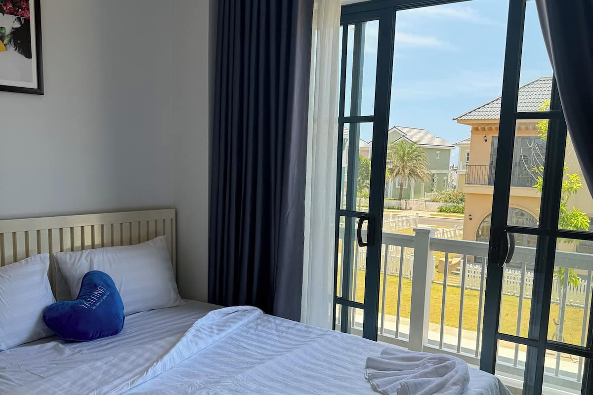 A hotel room with a bed, a heart-shaped pillow, a framed picture on the wall, and a view of the outside through a large window.