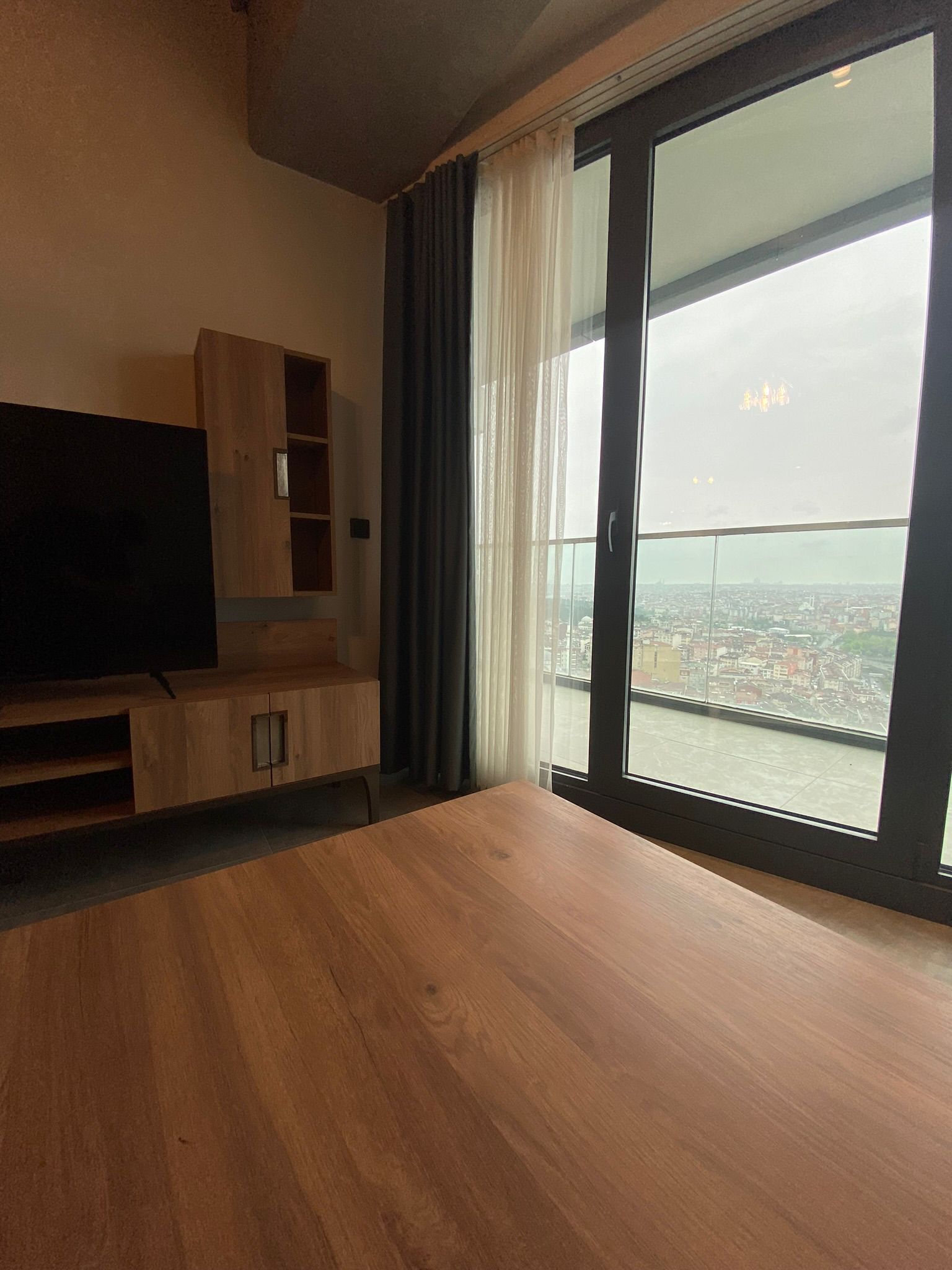 A modern living room with a large window, a flat-screen TV, wooden furniture, and a city view.