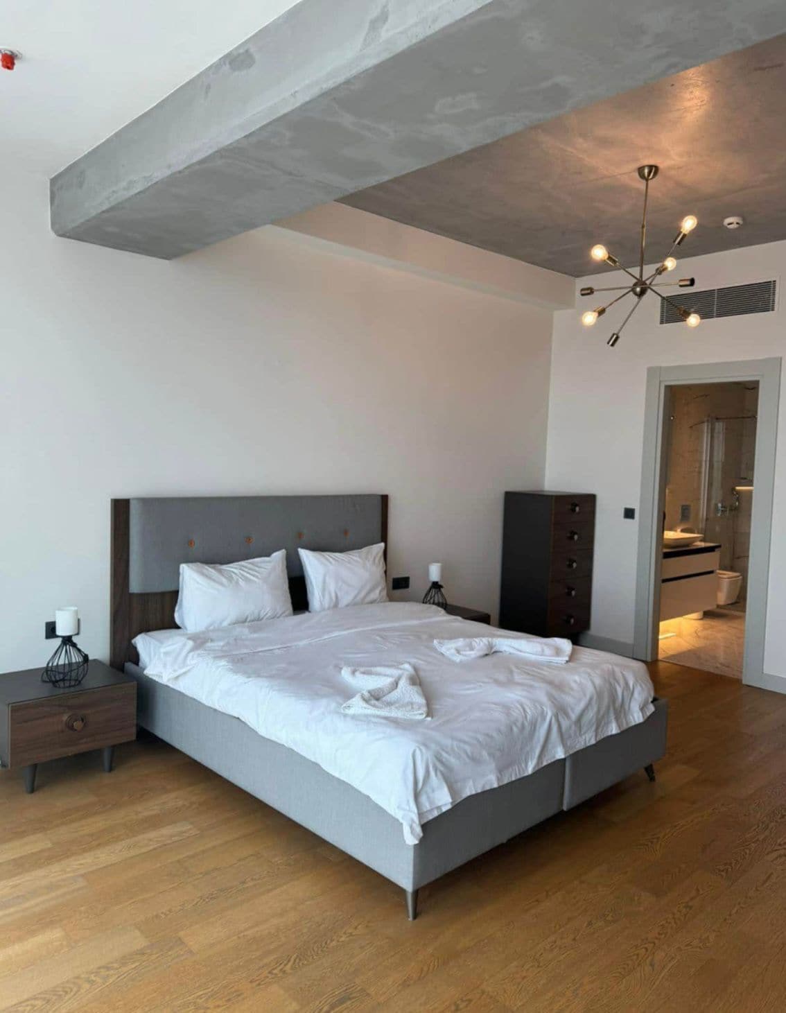 Quarto moderno com cama grande, mesa de cabeceira, cômoda e banheiro visível por uma porta aberta.