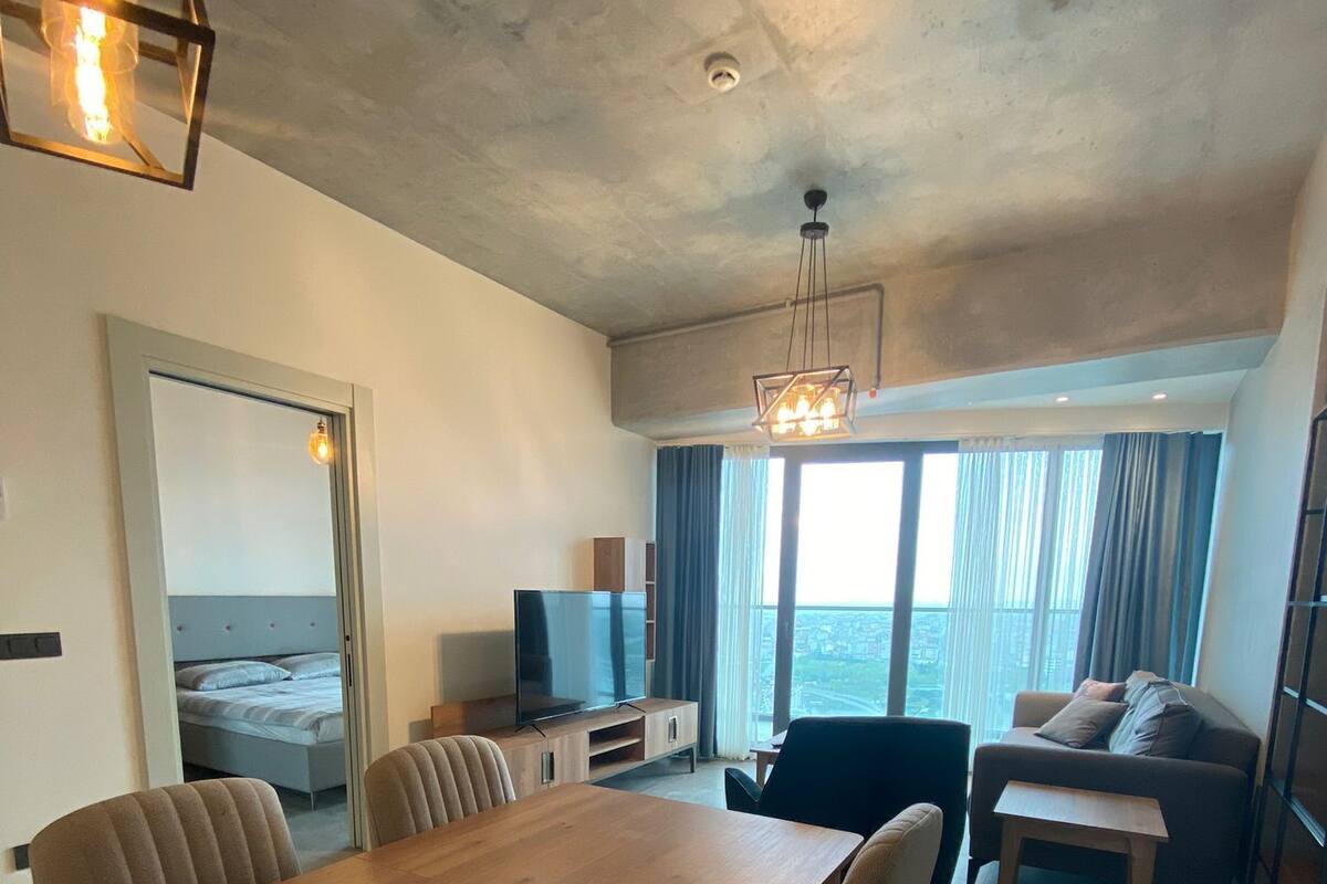Un salon moderne avec une table à manger en bois, des sièges confortables, un téléviseur et une grande fenêtre donnant sur la ville.