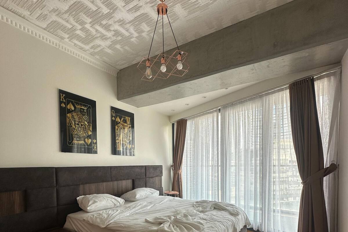 Une chambre moderne avec un grand lit, une tête de lit, un luminaire au plafond et une fenêtre avec des rideaux.