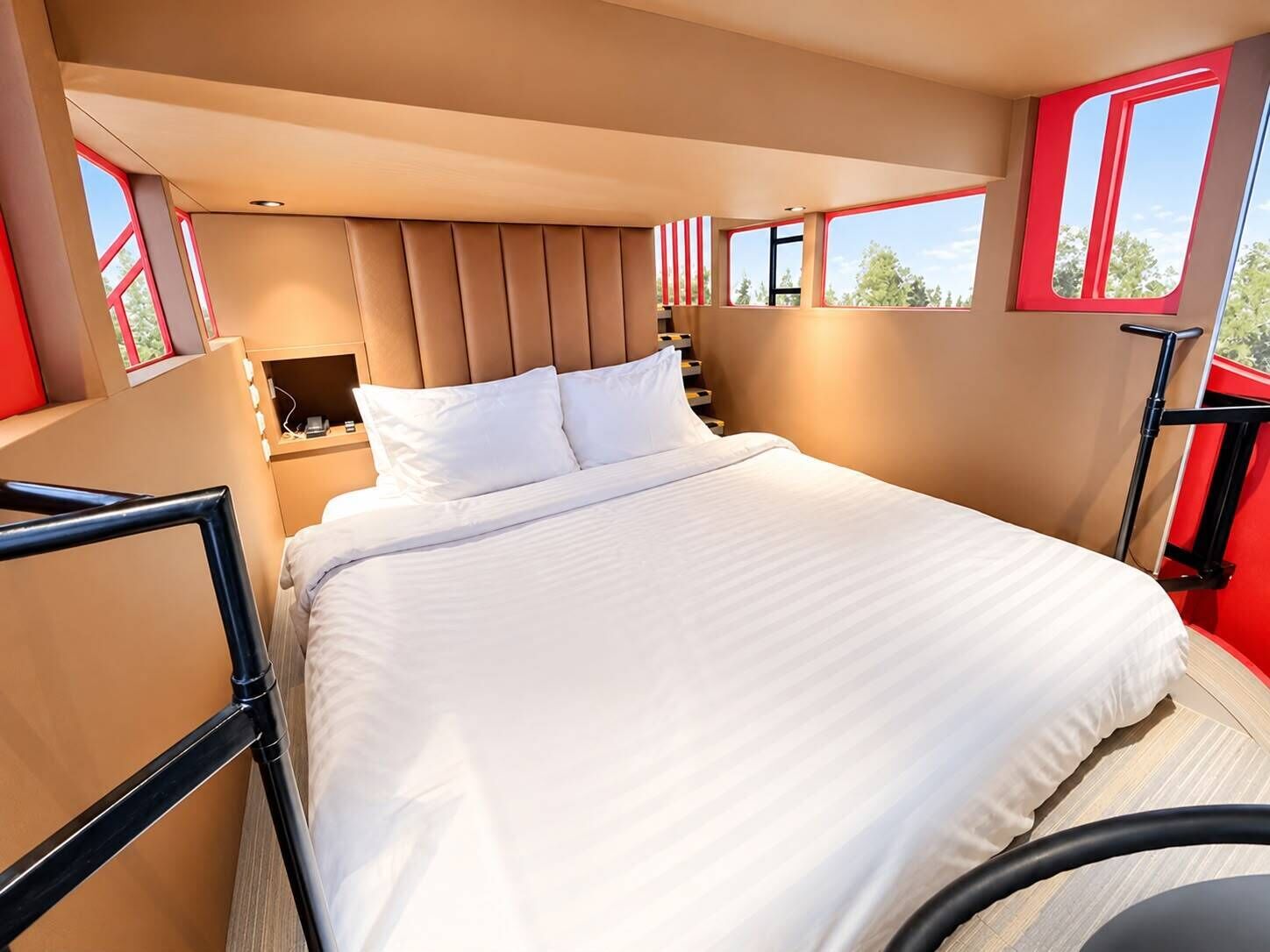 A modern hotel room with a large bed, a nightstand, a lamp, and a window with a red frame.