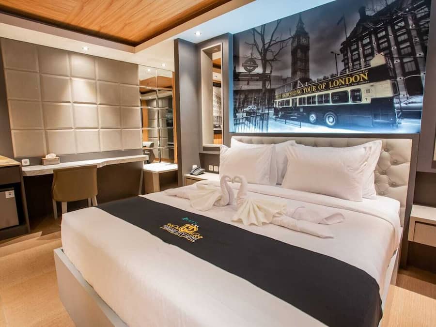 A hotel room with a large bed, a desk, and a chair. There is a poster of a London tour on the wall.