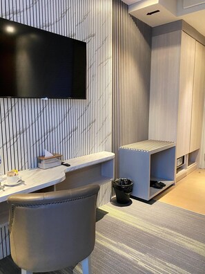 A modern hotel room with a flat-screen TV mounted on the wall, a desk with a chair, and a small seating area with a bench.