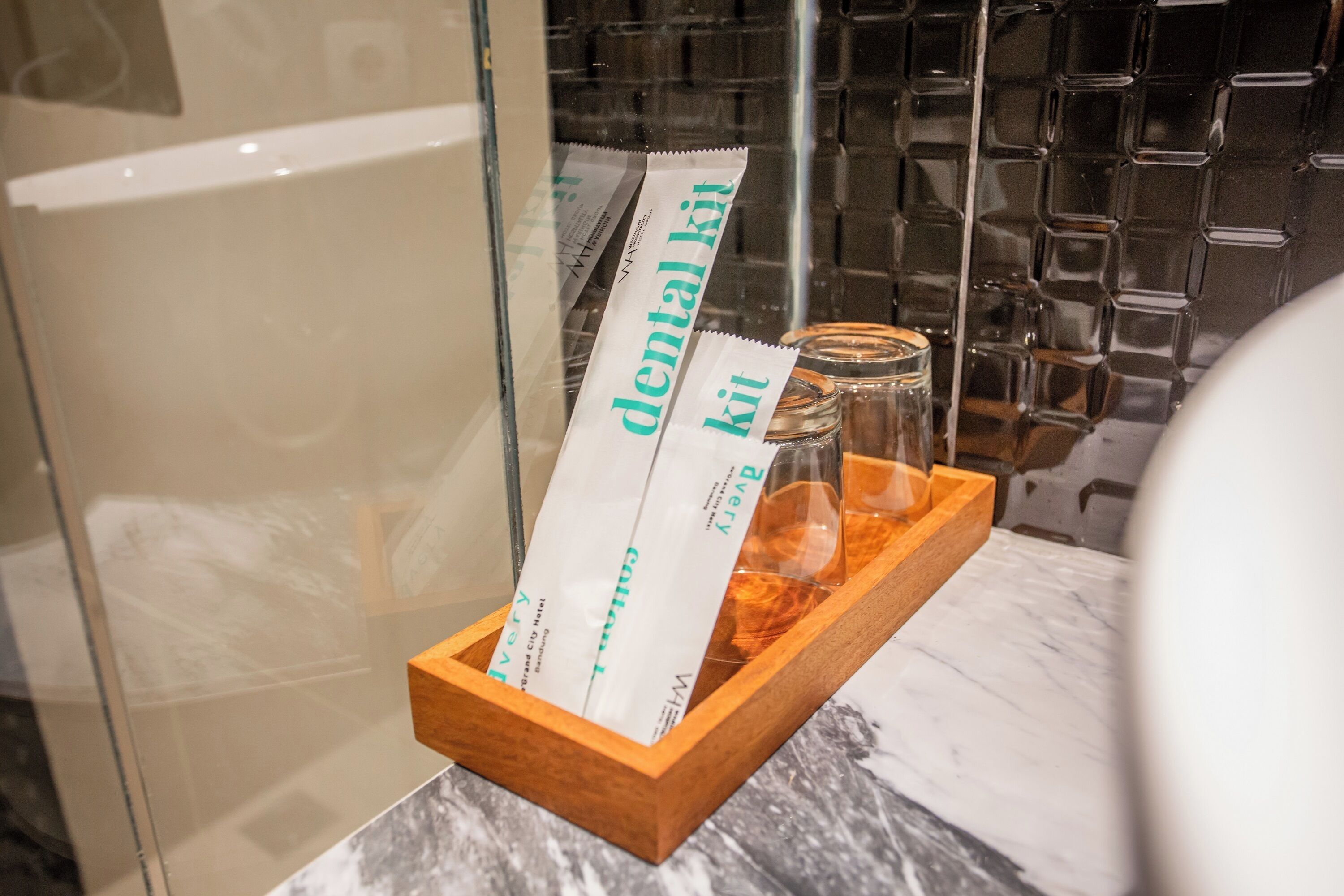 A wooden tray with dental kits and a glass jar in a bathroom with a marble countertop and a glass shower enclosure.