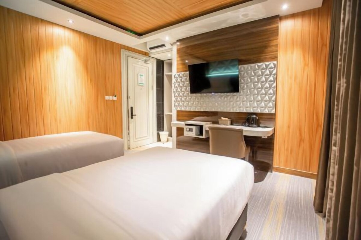 A hotel room with two beds, a desk, a TV mounted on a textured wall, and wooden paneling.