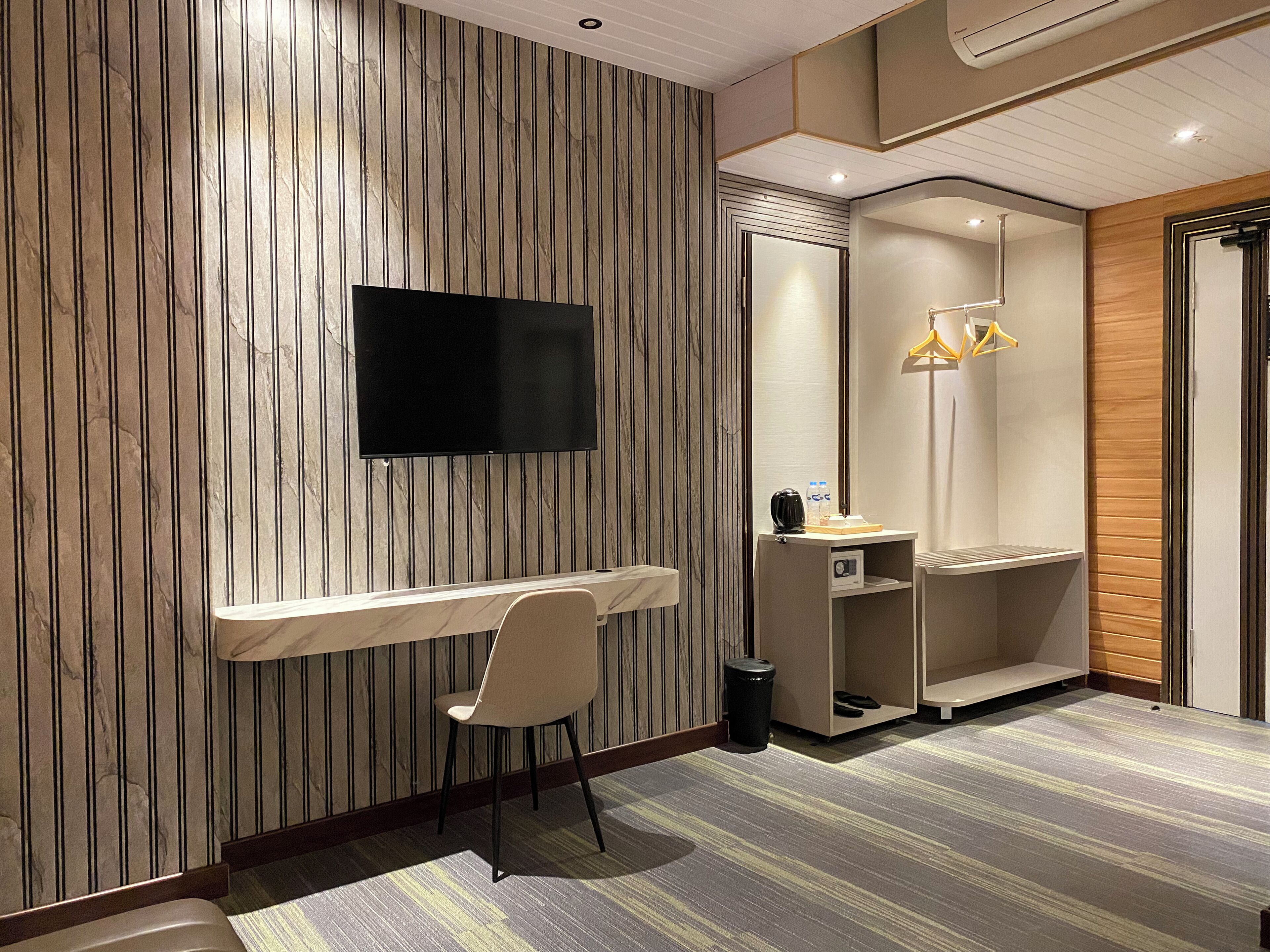 Ein modernes Hotelzimmer mit Flachbildfernseher an einer Wand aus Holzpaneelen, einem Schreibtisch mit Stuhl, einem Schminktisch mit Spiegel und einer Duschkabine mit Handtuchhalter.