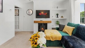 Un saló amb una llar de foc, un sofà verd, una televisió que mostra 'NETFLIX' i un ram de flors.