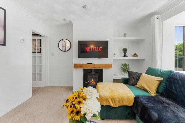 Ein Wohnzimmer mit Kamin, einem grünen Sofa, einem Fernseher, der 'NETFLIX' anzeigt, und einem Blumenstrauß.