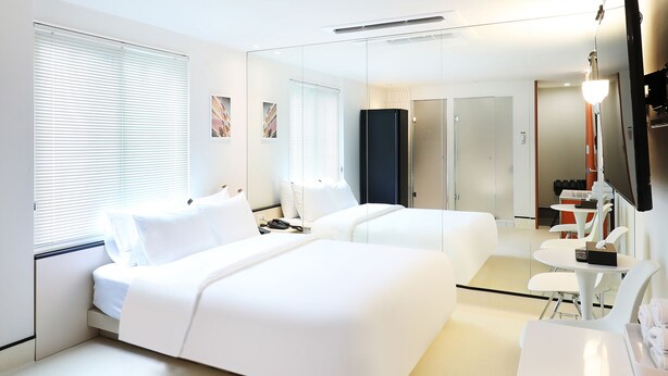 Une chambre d’hôtel moderne équipée de deux lits, d’un bureau, d’une chaise, d’une télévision et d’une salle de bain visible à travers une paroi en verre.