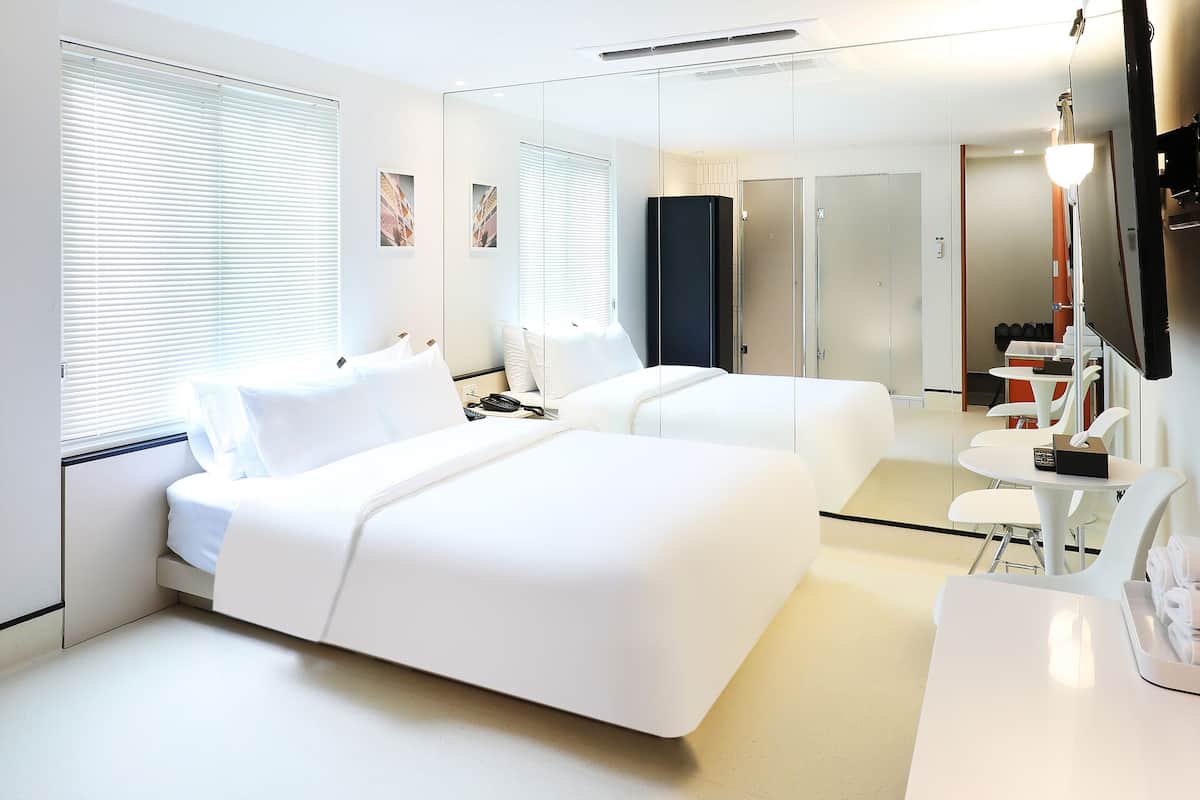 Une chambre d’hôtel moderne équipée de deux lits, d’un bureau, d’une chaise, d’une télévision et d’une salle de bain visible à travers une paroi en verre.