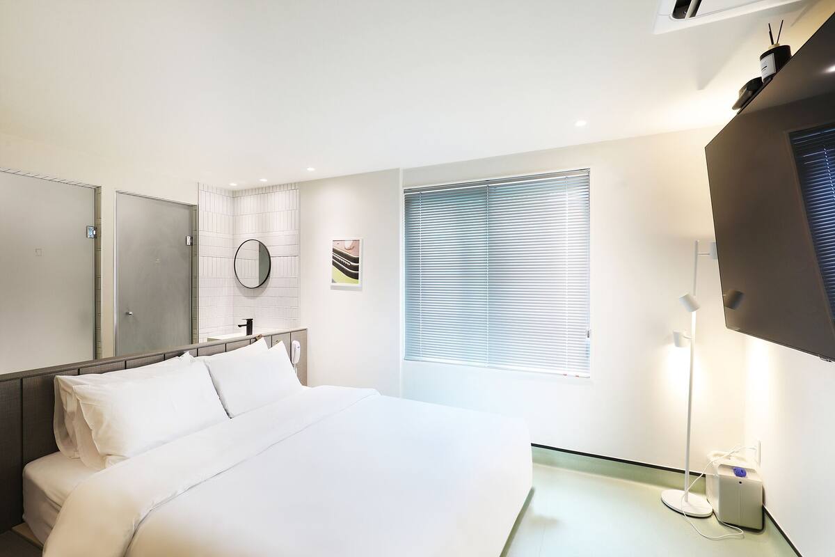 Une chambre d’hôtel comprenant un lit, une table de chevet, un miroir, une fenêtre avec des stores et un téléviseur fixé au mur.