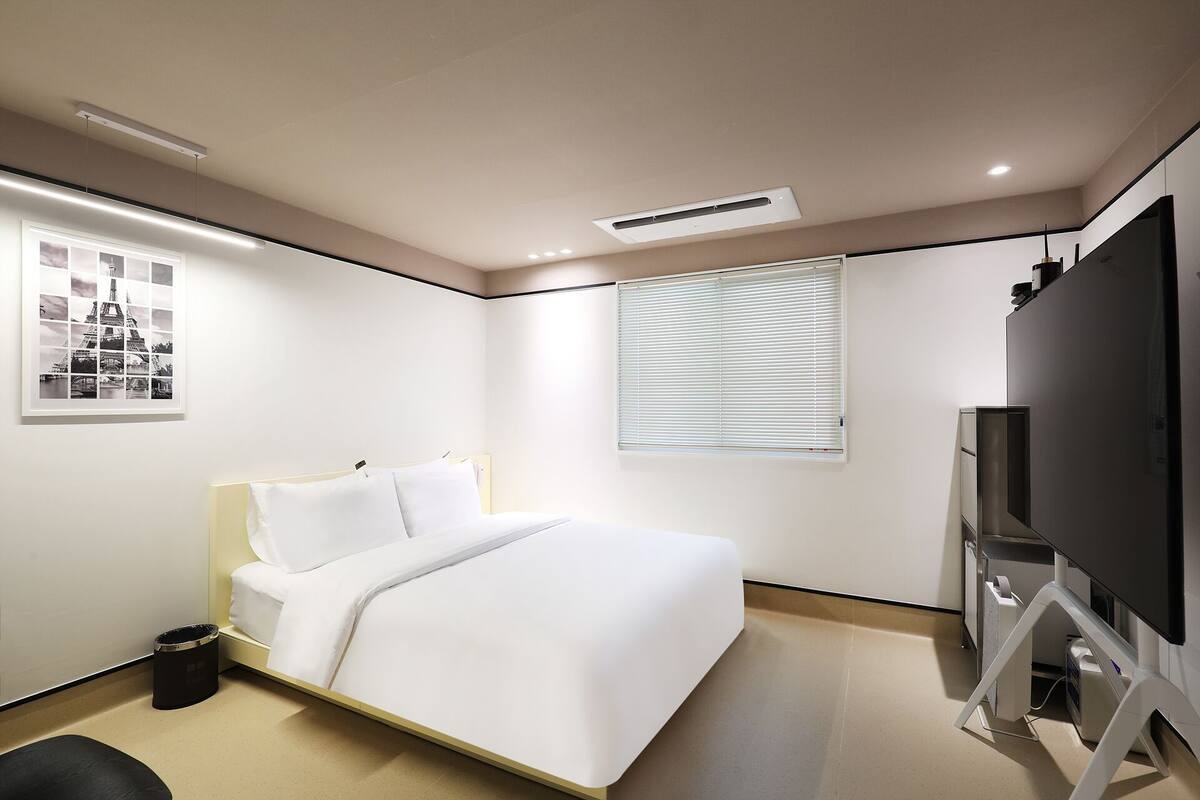 Une chambre d’hôtel moderne équipée d’un lit, d’une télévision, d’une fenêtre avec stores et d’un tableau représentant la Tour Eiffel au mur.