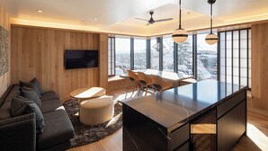 Uma sala de estar moderna com uma TV grande, um sofá, uma área de refeições com cadeiras e vista para uma paisagem nevada através de grandes janelas.