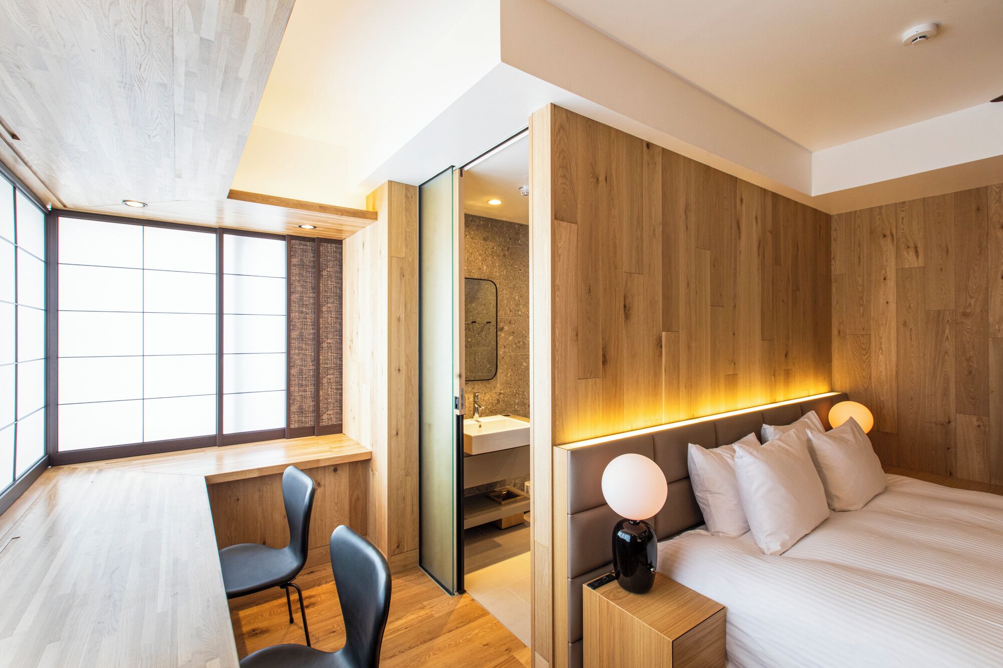 A modern hotel room with a wooden floor, a desk with two chairs, a large bed with a headboard, a bedside table with a lamp, and a large window with a view.