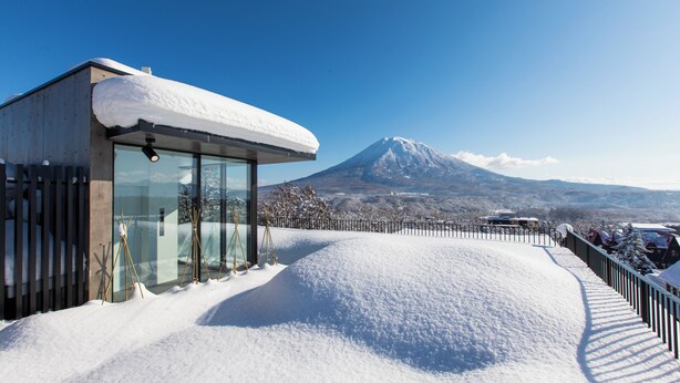 Um edifício moderno com grandes janelas de vidro, telhado coberto de neve e vista para uma montanha nevada ao fundo.