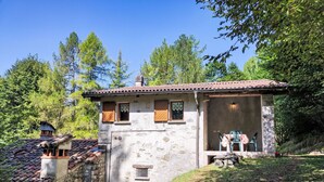 A rustic stone house with a tiled roof, a small porch with chairs, and a stone wall. - Vacation home Relax by Interhome (Carlazzo)