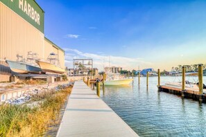 A waterfront walkway leading to a dock with boats, a large building labeled "HARBOR" on the left, and a clear sky.