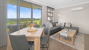 A modern living room with a dining area, large windows, a grey sofa, and a wooden coffee table. - Luxurious 2-Bedroom Spa Apartment with Ocean Views (KRONKUP)