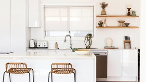 A modern kitchen with white cabinetry, a central island with two wicker chairs, a built-in dishwasher, and wooden shelves above the sink. - Casa Blanca - Lakeside Luxury (Mount Warrigal)