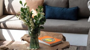 Un salón con un sofá, una mesa de centro con un libro y un jarrón de flores, y el pie de una mesa de madera.