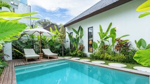 A swimming pool with two lounge chairs and a sun umbrella, surrounded by lush greenery and a white building with a tiled roof.