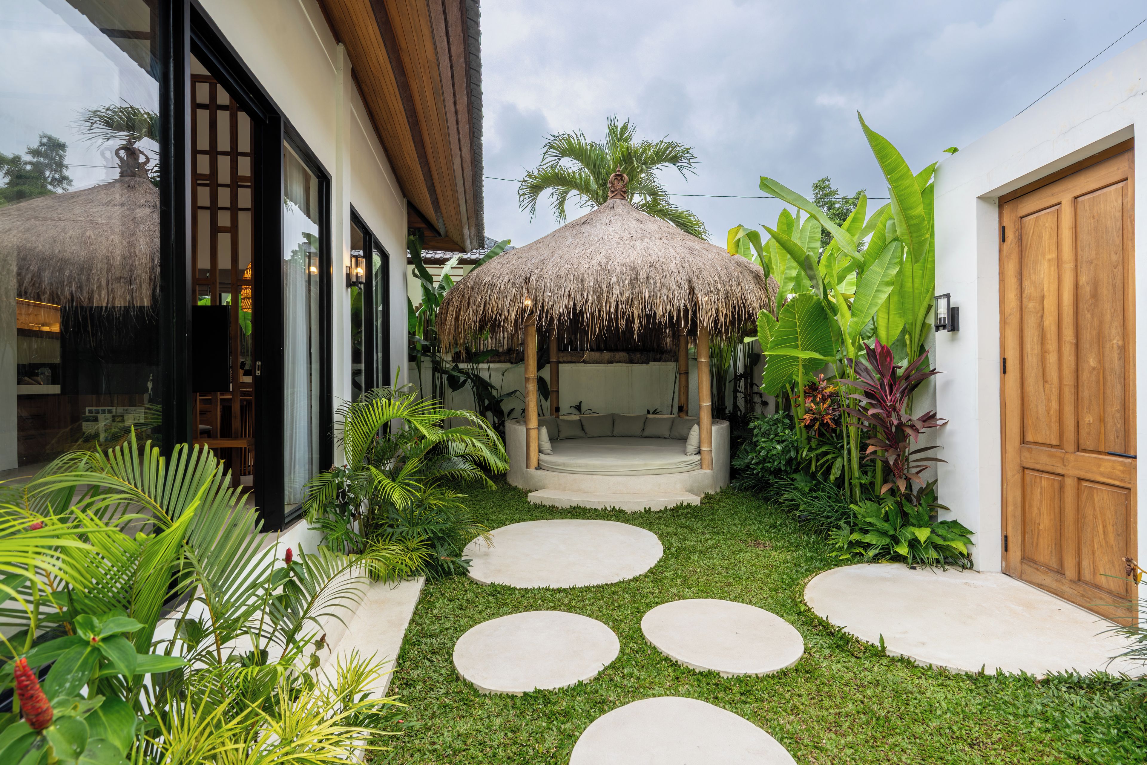 Um jardim tropical com um gazebo de palha, uma porta de madeira e um caminho de concreto.