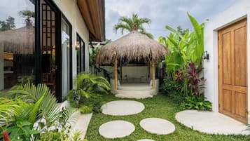 Um jardim tropical com um gazebo de palha, uma porta de madeira e um caminho de concreto.
