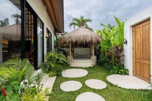 A tropical garden with a thatched gazebo, a wooden door, and a concrete pathway. - Alpha Villa by DH (Ubud)