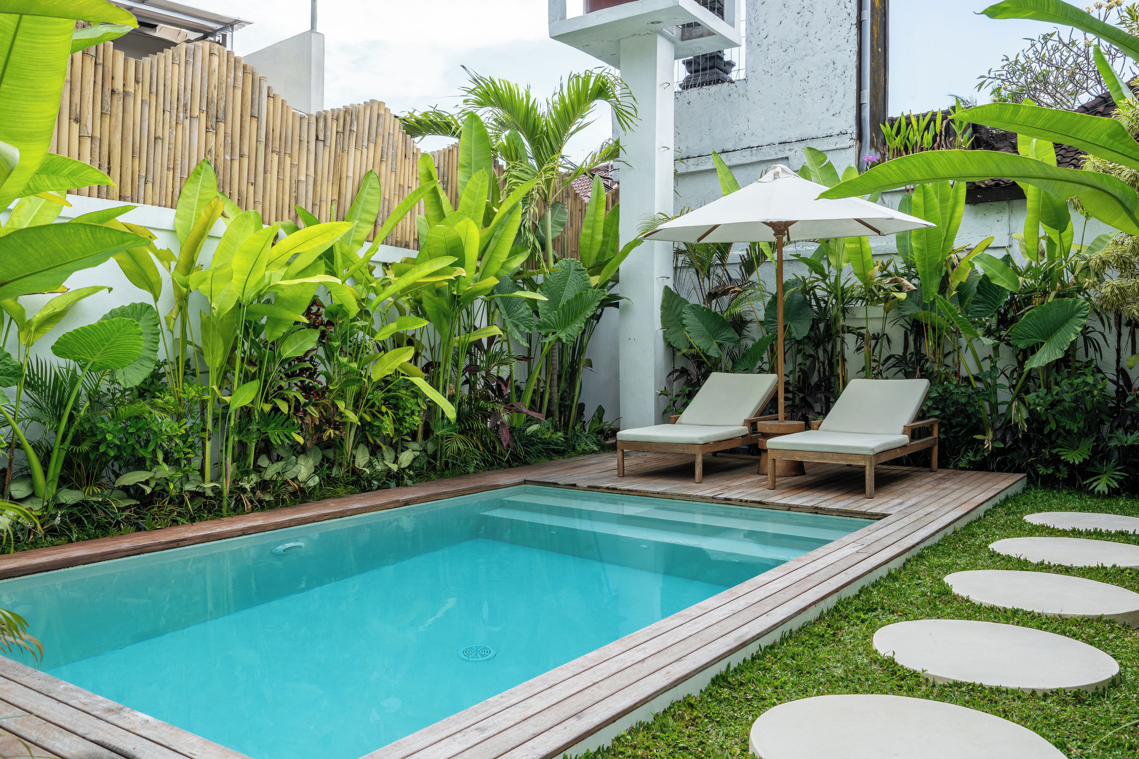 Una piscina rectangular con cubierta de madera, rodeada de abundante vegetación y una valla blanca.