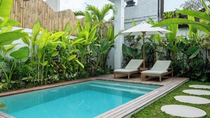 A rectangular swimming pool with a wooden deck, surrounded by lush greenery and a white fence. - Alpha Villa by DH (Ubud)