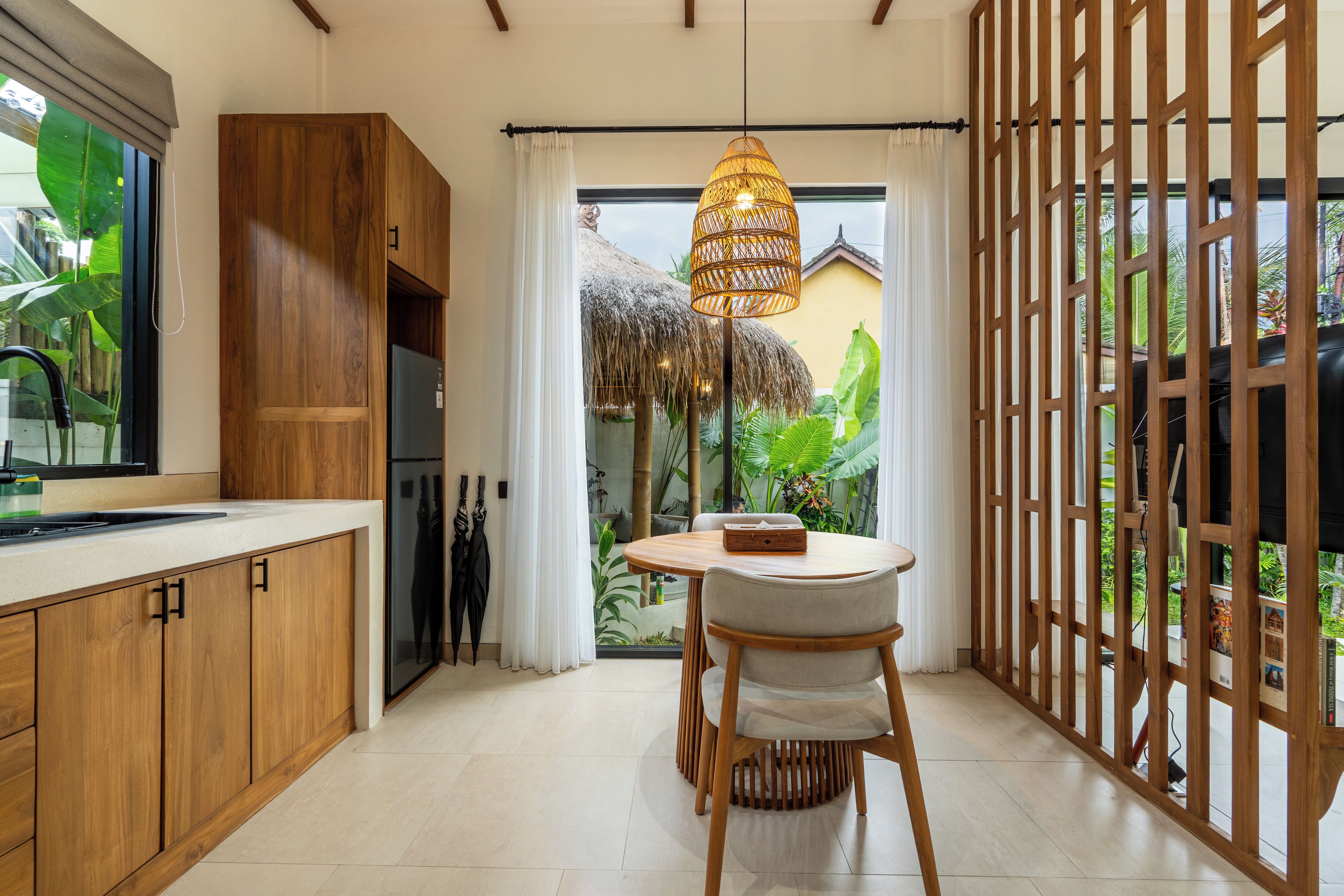 A modern kitchen with wooden cabinets, a dining area with a table and chairs, and a view of a tropical garden through a glass door.