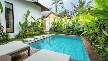 Piscina com deck de madeira e espreguiçadeiras, cercada por vegetação tropical exuberante e um edifício branco com janela.