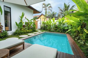 A swimming pool with wooden decking and lounge chairs, surrounded by lush tropical plants and a white building with a window. - Alpha Villa by DH (Ubud)