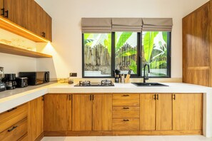 A modern kitchen with wooden cabinets, a white countertop, and a window with a view of greenery. - Alpha Villa by DH (Ubud)