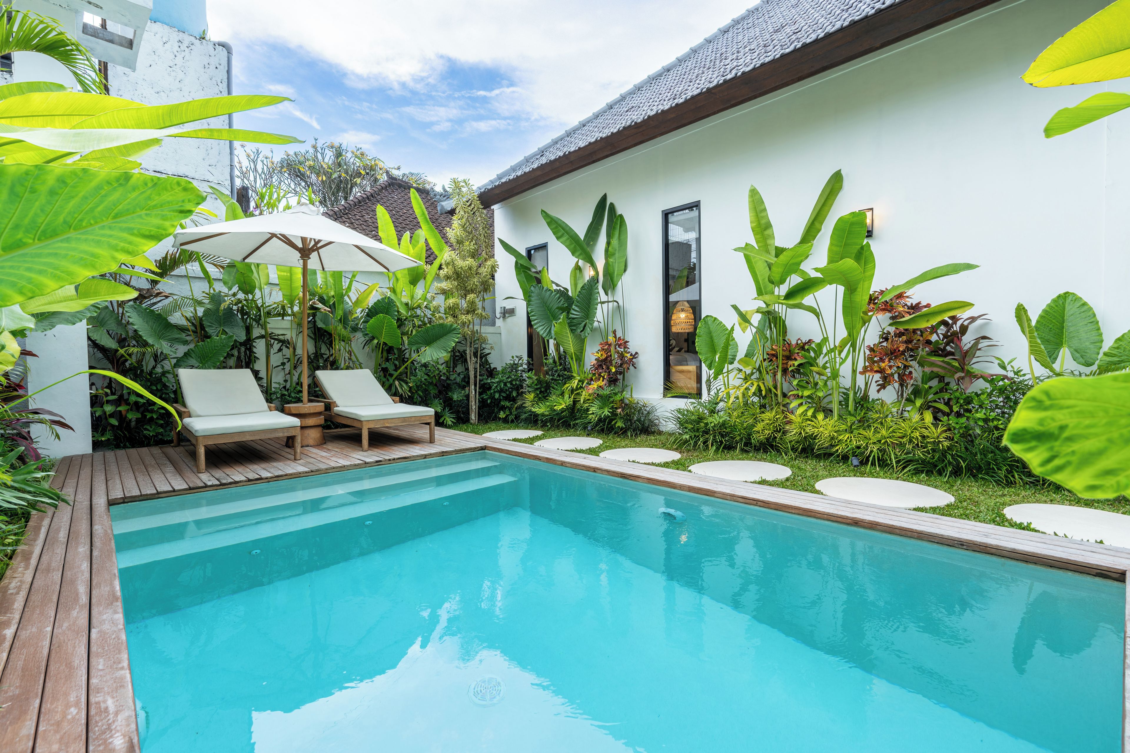 A swimming pool with two lounge chairs and a sun umbrella, surrounded by lush greenery and a white building with a tiled roof.