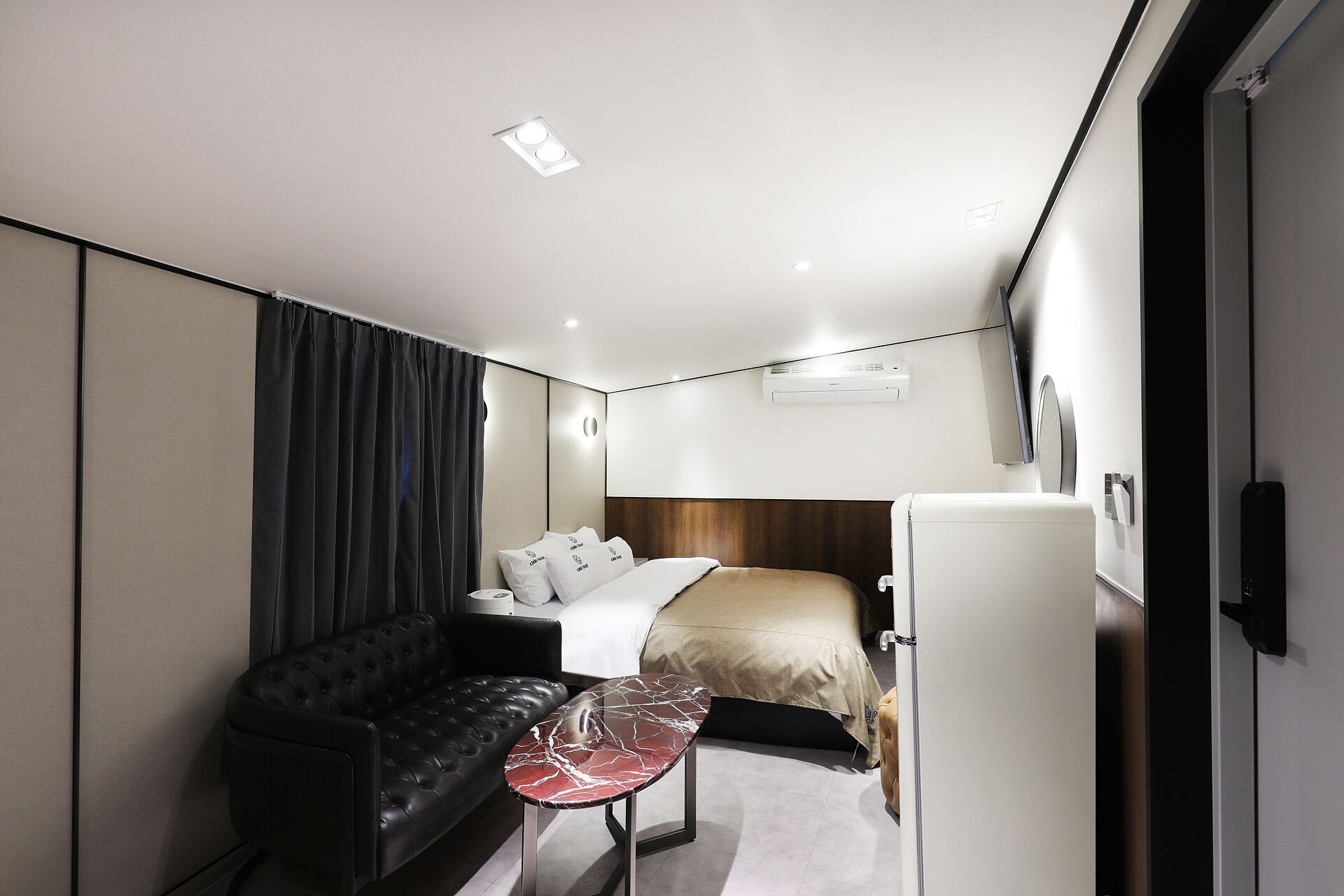 A compact hotel room with a bed, a sofa, a small table, and a refrigerator.