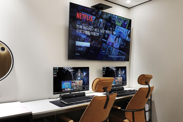 A modern office space with a large wall-mounted screen displaying a Netflix interface, two desktop computers, and two brown chairs. - Cielo Hotel (Jecheon)
