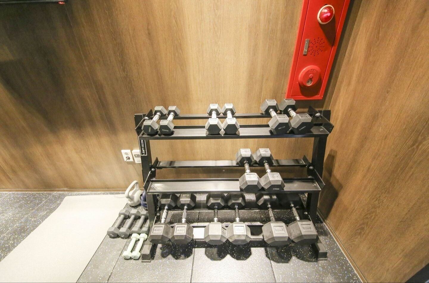 Un juego de pesas ordenado en un soporte dentro de una habitación con paredes de madera y un botón de emergencia rojo.