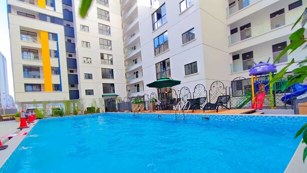 Une piscine entourée de chaises longues et de parasols, elle-même bordée d’un immeuble de grande hauteur doté de balcons.