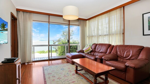 A spacious living room with a leather sofa, a wooden coffee table, and a large window offering a view of the sea. - Jimmys Beach Waterfront (Hawks Nest)