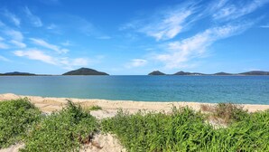 A beach with clear blue water, distant islands, and green vegetation in the foreground. - Jimmys Beach Waterfront (Hawks Nest)