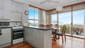 A modern kitchen with a central island, dining area with chairs, a clock, and a view of the sea. - Jimmys Beach Waterfront (Hawks Nest)